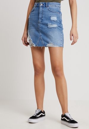 Mini-jupe en jean avec des zones usées et un ourlet effiloché portée avec des baskets montantes noires et blanches sur une personne debout.