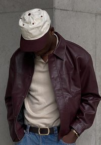 Veste en cuir bordeaux, polo beige à côtes, jeans bleus, casquette blanche avec visière bordeaux, design décontracté, couture détaillée, accessoires en métal minimalistes.