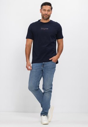 Uomo in piedi contro uno sfondo bianco, indossa una maglietta navy Tommy Hilfiger, jeans blu e scarpe da ginnastica bianche, con una mano in tasca.