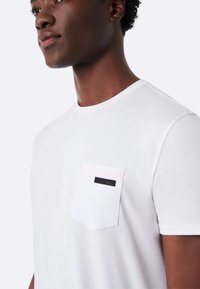 T-shirt bianco a girocollo realizzata in cotone, con una tasca bordata di rosa e un'etichetta nera sul lato sinistro del petto. Tessuto morbido, maniche corte.