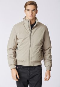 Giacca bomber beige con colletto alto, chiusura con zip e polsini a coste. Presenta tasche laterali e orlo dritto. Indossata sopra una maglietta nera.