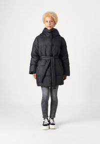 Giacca a vento nera con cappuccio, vita con cintura e design trapuntato; abbinata a jeans skinny grigi e sneakers nere con suola bianca.