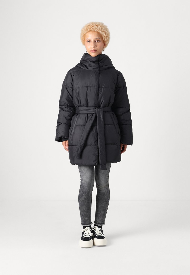 Giacca a vento nera con cappuccio, vita con cintura e design trapuntato; abbinata a jeans skinny grigi e sneakers nere con suola bianca.