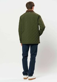 Homme portant une veste matelassée vert olive, un pantalon bleu marine et des chaussures blanches, debout dos à la caméra sur un fond uni.