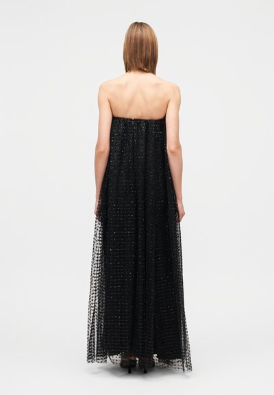 Woman standing facing away wearing a strapless black floor-length dress with a textured overlay and high-heeled shoes on a white background.