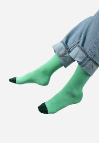 Zielone skarpetki z gładkiego materiału, z ciemnozielonym palcem. Noszone z podwiniętymi niebieskimi dżinsami, prezentującymi luźny krój i design.