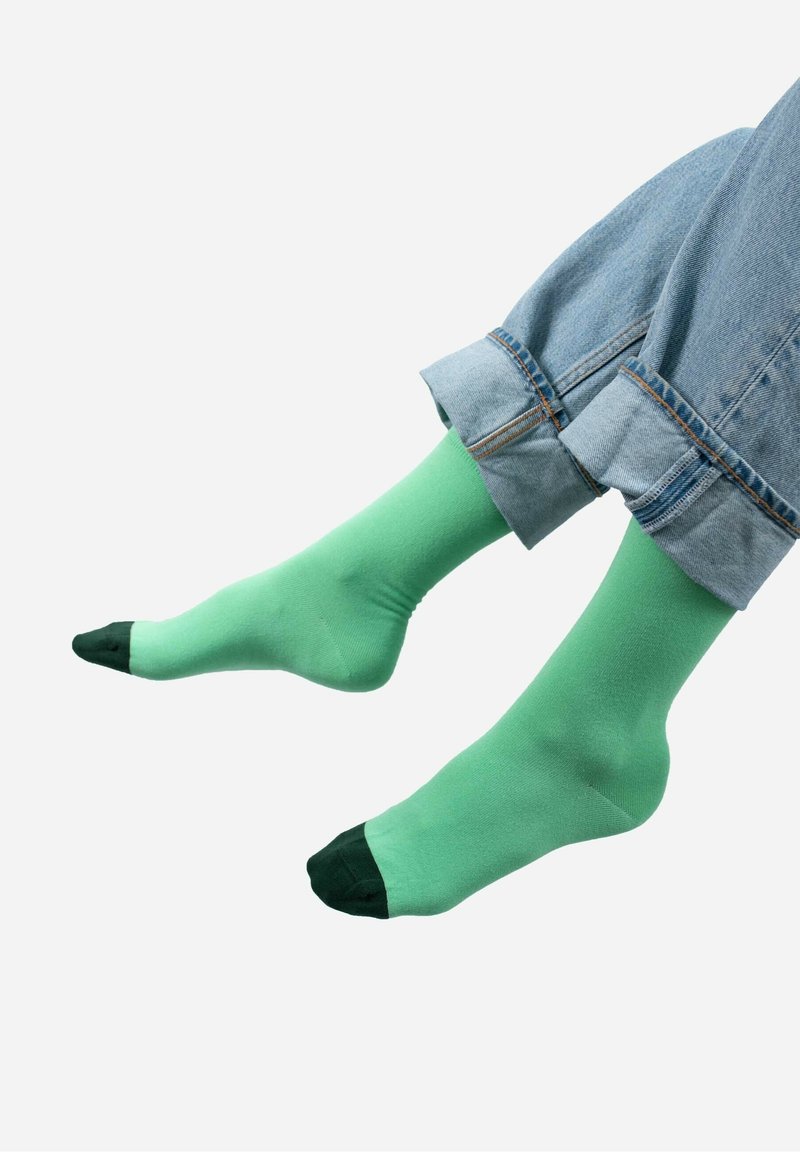 Zielone skarpetki z gładkiego materiału, z ciemnozielonym palcem. Noszone z podwiniętymi niebieskimi dżinsami, prezentującymi luźny krój i design.