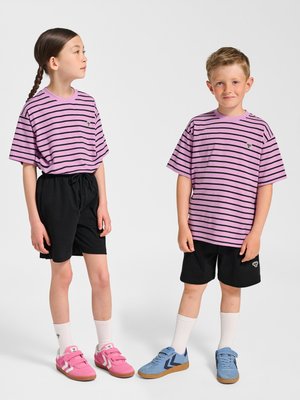 Zwei Kinder tragen passende lila-schwarz gestreifte Shirts und schwarze Shorts, eines mit pinken Schuhen, das andere mit blauen Schuhen, und stehen nebeneinander.