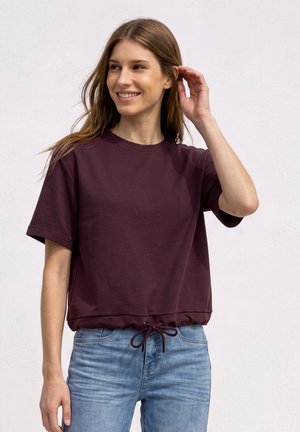 Ung kvinde smiler, iført en mørk lilla t-shirt med korte ærmer og snørrehæm samt lyseblå jeans, mod en lys baggrund.