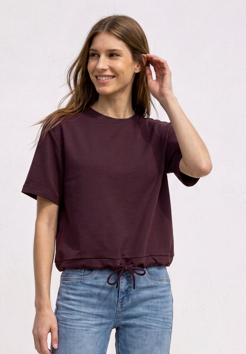 Jeune femme souriante, portant un t-shirt violet foncé à manches courtes avec un cordon à l'ourlet et un jean bleu clair, sur un fond clair.