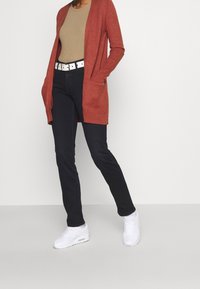 Roestkleurige lange cardigan met zakken over een beige top, gecombineerd met donkere jeans en witte sneakers. Eenvoudige, casual outfit.