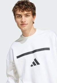 Biely sveter s okrúhlym výstrihom, ktorý má čierny horizontálny pruh a logo Adidas. Vyrobený z mäkkého materiálu, s voľným strihom.