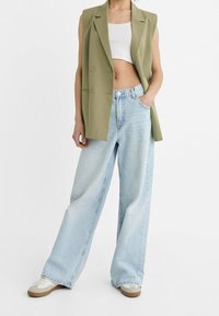 Blazer sans manches vert sur un crop top blanc, associé à un jean large bleu clair et des baskets beiges. Tissu texturé et coupe ample.