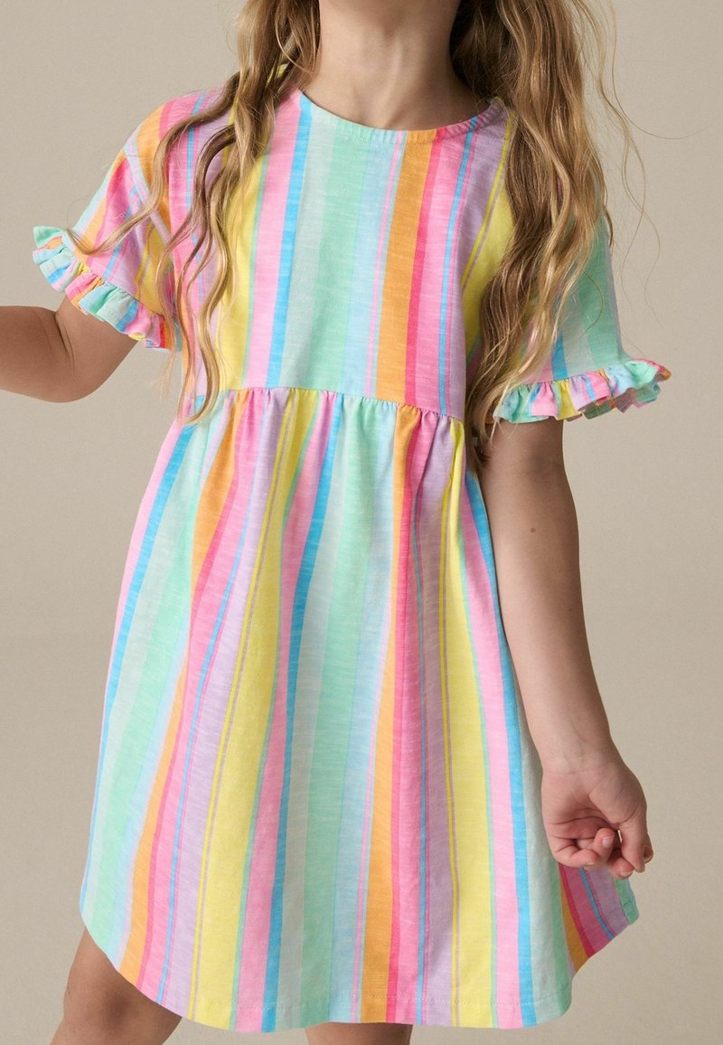 Jeune fille portant une robe à manches courtes avec des rayures verticales pastel et des bords de manches à volants, debout devant un fond uni.