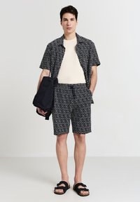 Set di camicia e pantaloni corti a disegno geometrico in bianco e nero, realizzato in tessuto leggero. Sandali neri e borsa scura.