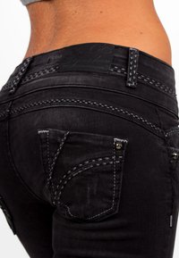 Jeans in denim nero con cuciture bianche, caratterizzati da tasche posteriori decorative e un'etichetta in pelle sulla vita. Inquadratura ravvicinata.