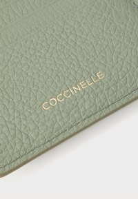 Portefeuille en cuir texturé vert avec le nom de marque embossé "COCCINELLE" en or, doté de fentes pour cartes et d'un design épuré et minimaliste.