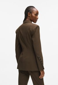 Brauner taillierter Blazer mit figurbetontem Schnitt, scharfen Revers und sechs schwarzen Knöpfen an den Manschetten, zeigt strukturierte Nähte und eine glatte Textur.