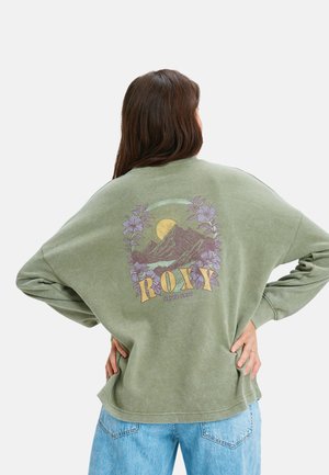 Femme avec de longs cheveux bruns portant un t-shirt à manches longues vert avec une montagne, un soleil, des fleurs violettes, et le texte "ROXY" dans le dos, associé à un jean bleu.
