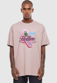 Lyserød t-shirt med korte ærmer, der har et grafik af en lyserød mobiltelefon og dekore tekst, der siger "Hotline" med legende akcenter.