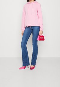 Felpa rosa con cappuccio e lacci, jeans a zampa d'elefante blu e tacchi alti rosa brillante con dettagli a forma di cuore. Borsa a tracolla rosa in mano.