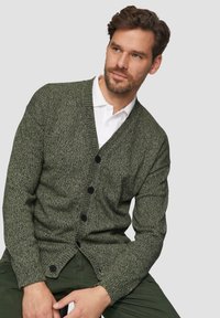 Grüne Strickjacke mit strukturiertem Muster, V-Ausschnitt und schwarzen Knöpfen. Kombiniert mit einem weißen Poloshirt und eng anliegenden olivfarbenen Hosen.