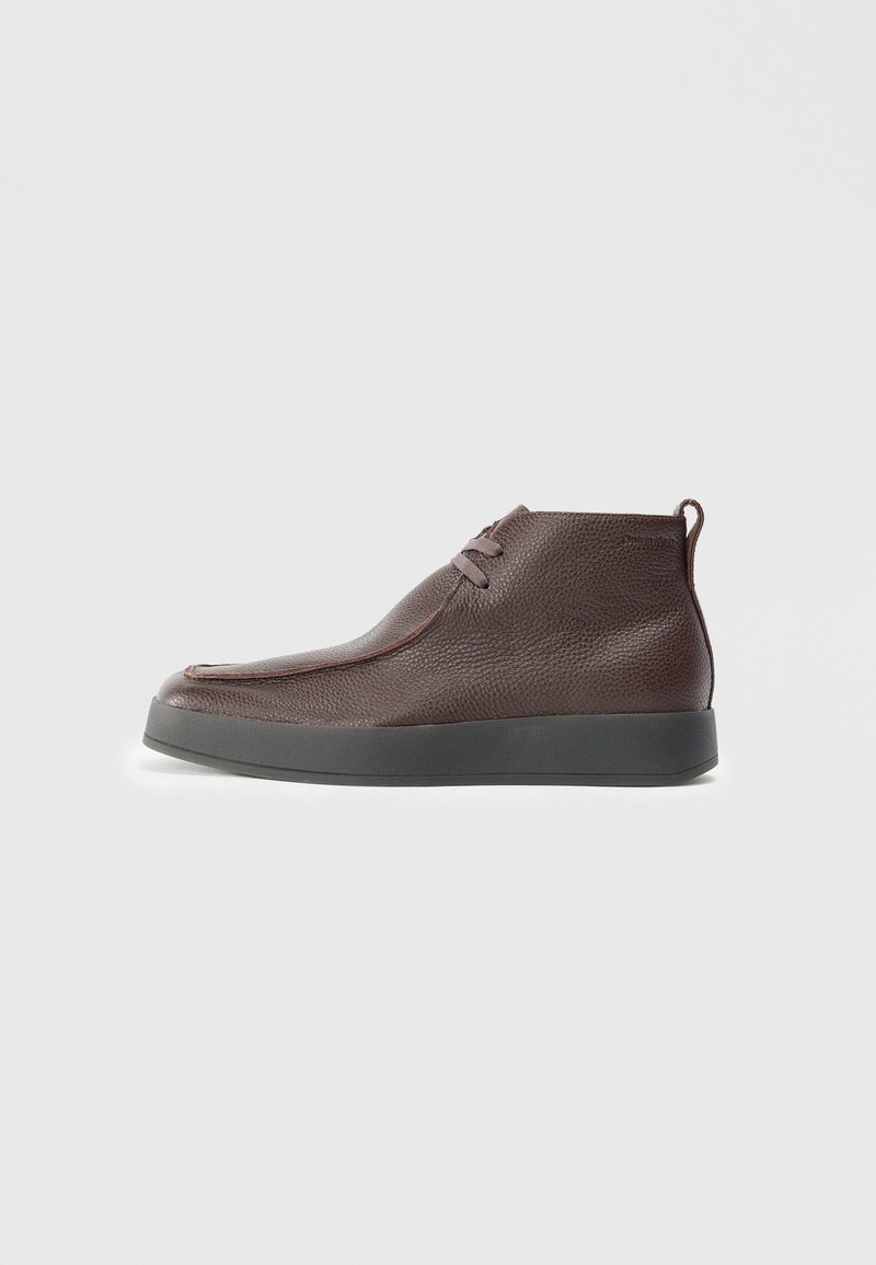 Botas de cuero marrón hasta el tobillo con una superficie texturizada, punta redondeada suave y suela de goma gris, que presentan dos ojales para los cordones.