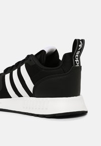 Svart idrottssko med mesh-övre och läderdetaljer. Har tre vita horisontella ränder och en gummisula med en texturerad häl.