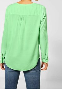 Grön långärmad blus med rundad halsringning, veckdetalj bak och knäppta manschetter, tillverkad av ett slätt tyg. Kombinerad med blå jeans.