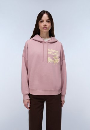 Frau trägt einen pinken Kapuzenpullover mit gelbem Aufdruck und dunkelbraune Hose, steht vor einem einfarbig hellgrauen Hintergrund.