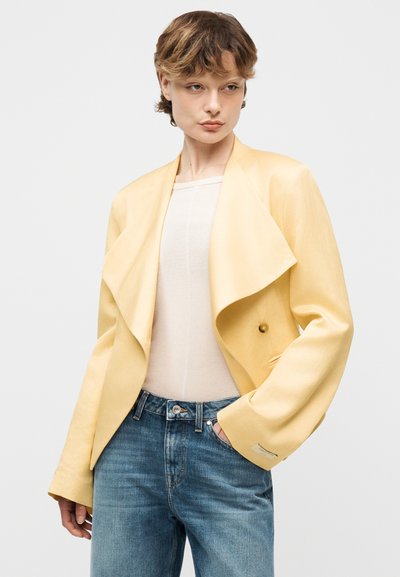 Νεαρό άτομο που φοράει ανοιχτό κίτρινο φαρδύ σακάκι, λευκό τοπ και μπλε τζιν, στέκεται με το ένα χέρι στην τσέπη καθώς ποζάρει σε απλό φόντο.