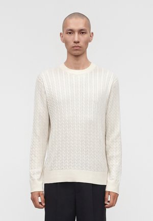 Mężczyzna z ogoloną głową, ubrany w biały sweter warkoczowy i czarne spodnie, stojący na tle jednolitego jasnego tła.