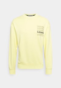 Jasnożółta długozasłonowa bluza z rękawami, z prążkowanymi mankietami i dolnym brzegiem, z powtarzającym się napisem "Lacoste" w konturze i pogrubieniu na piersi.