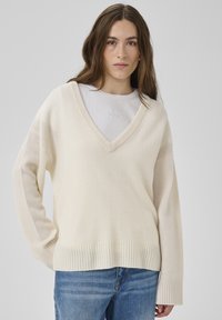 Pull crème col en V avec manches longues et larges et poignets côtelés. Présente une texture douce et une coupe ample, superposé sur un t-shirt blanc.