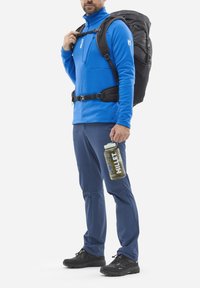Mann in blauer Outdoor-Jacke und Hose, mit einem schwarzen Wanderrucksack und einer Wasserflasche mit Griff und Markenlogo.