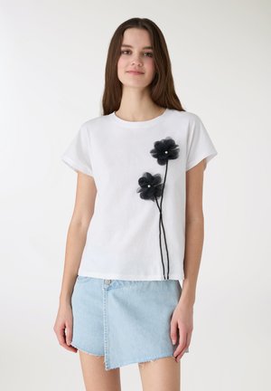 Biały t-shirt z krótkimi rękawami, ozdobiony dwoma czarnymi przezroczystymi aplikacjami kwiatowymi z perłowymi akcentami na czarnym łodydze. Noszony z dżinsową spódnicą.