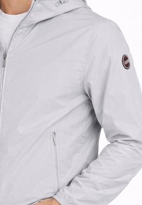 Férfi világosszürke cipzáras kapucnis dzsekiben, cipzáras oldalzsebbel és kerek, piros-fekete logófolttal a felső ujján.