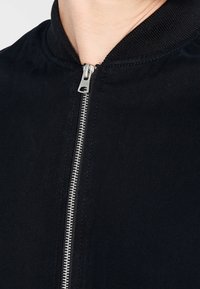 Close-up of a zipped black jacket with a silver metal zipper and ribbed collar, showing part of a person’s neck and chin.