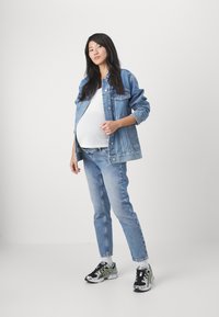 Chaqueta de denim, camiseta blanca, jeans de un azul claro y zapatillas negras y verdes. La modelo está de pie con una mano apoyada en su abdomen.
