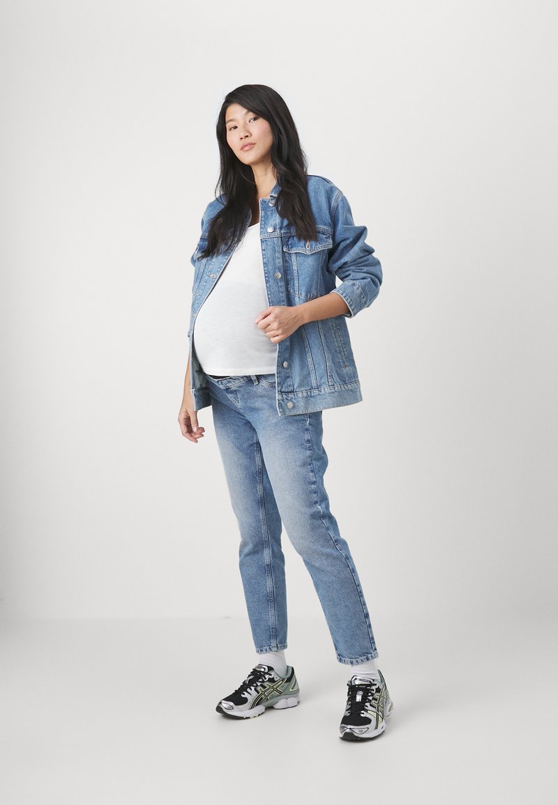 Chaqueta de denim, camiseta blanca, jeans de un azul claro y zapatillas negras y verdes. La modelo está de pie con una mano apoyada en su abdomen.