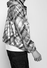 Silverprickigt jacka med huva, med en glänsande textur och svarta ränder. Matchad med en vit skjorta och svarta cargo-byxor.