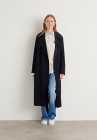 Abrigo largo negro con un cuello amplio, superpuesto a un suéter ligero y una camisa a rayas, combinado con unos vaqueros de mezclilla azul claros y zapatillas blancas.