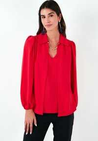 Red blouse with a buttoned collar, puff sleeves, and a smooth texture. Worn over a matching sleeveless top, paired with black pants.