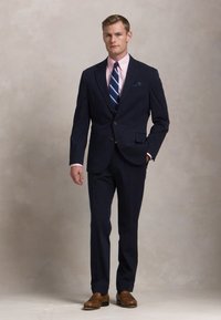 Mann steht da und trägt einen marineblauen Nadelstreifenanzug, ein rosa Hemd, eine blau gestreifte Krawatte, braune Schuhe und ein Einstecktuch, Hintergrund neutral.