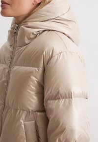 Beżowa kurtka puchowa z wysokim kołnierzem, kapturem i błyszczącym wykończeniem. Gruby pikowany wzór z bocznymi kieszeniami i zapięciem na zamek błyskawiczny.