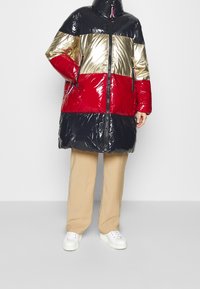 Puffjacka med glansiga svarta, guldiga och röda paneler, hög krage, dragkedjestängning och sidofickor. Bärs över beiga byxor och vita sneakers.