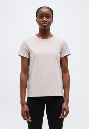 Žena s copánky, oblečená v světle béžovém tričku s krátkým rukávem a černých kalhotách, stojící na bílém pozadí.