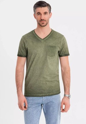 Camiseta verde oliva de cuello en V con un bolsillo, de mangas cortas con un puño en contraste, hecha de una tela texturizada. Llevada con jeans azul claro.