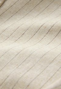 Tissu beige avec de fines rayures noires verticales et espacées régulièrement, montrant la texture et de légers plis.