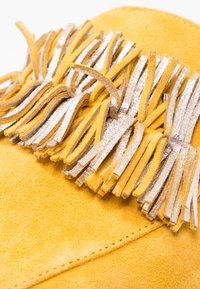 Mocassin en suédine jaune avec des détails en franges argentées et dorées sur le dessus. Texture douce avec des coutures visibles le long du bord.